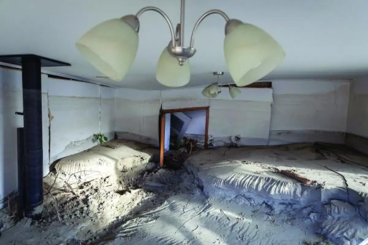 Damaged room with two ceiling lights, wallpaper shifted and floor covered with dirt and concrete