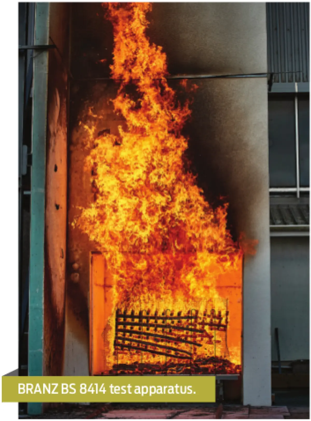 Photograph showing a vertical fire test apparatus with flames rising inside a tall enclosure, burning material positioned at the base