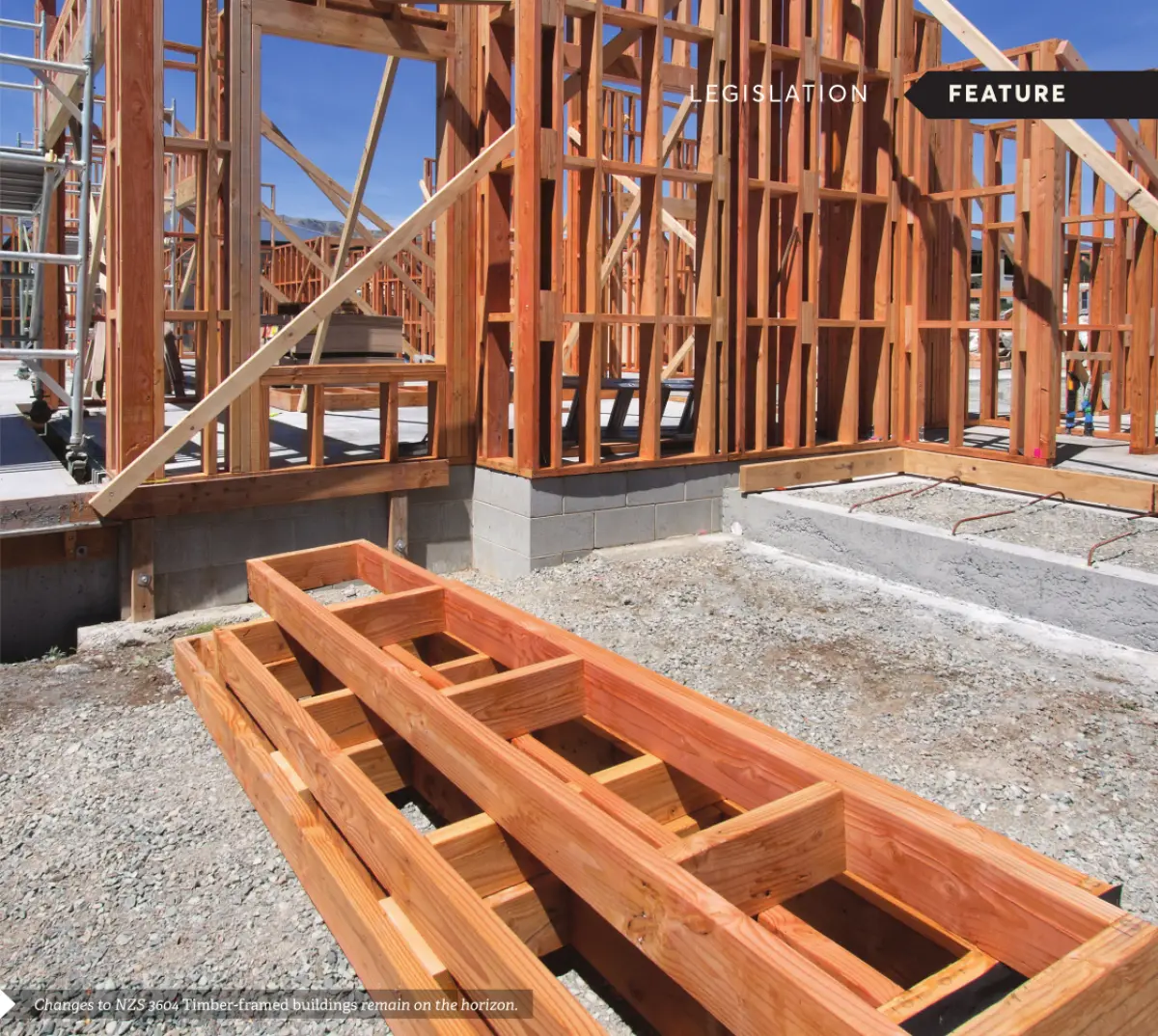 Photograph showing timber framing under construction on a concrete foundation, with stacked timber components in the foreground