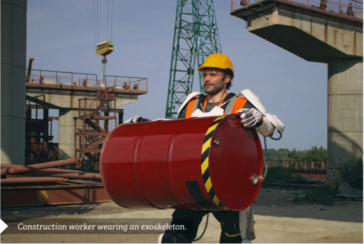Photograph showing a construction worker carrying a large barrel while wearing a powered exoskeleton at an industrial site