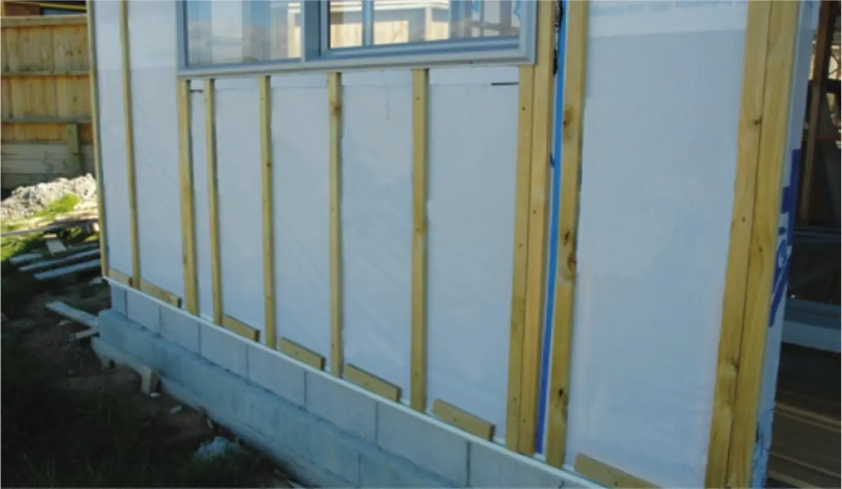 Photograph showing vertical timber battens fixed over a white wall surface, with a window above and a concrete foundation at the base