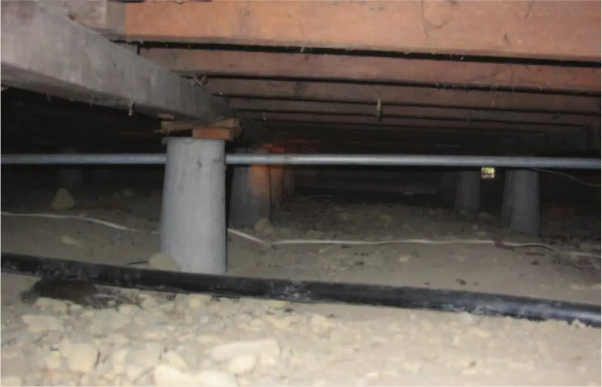 Photograph showing the underside of a timber house with concrete piles supporting floor framing above, bare ground below, and a horizontal pipe running between piles