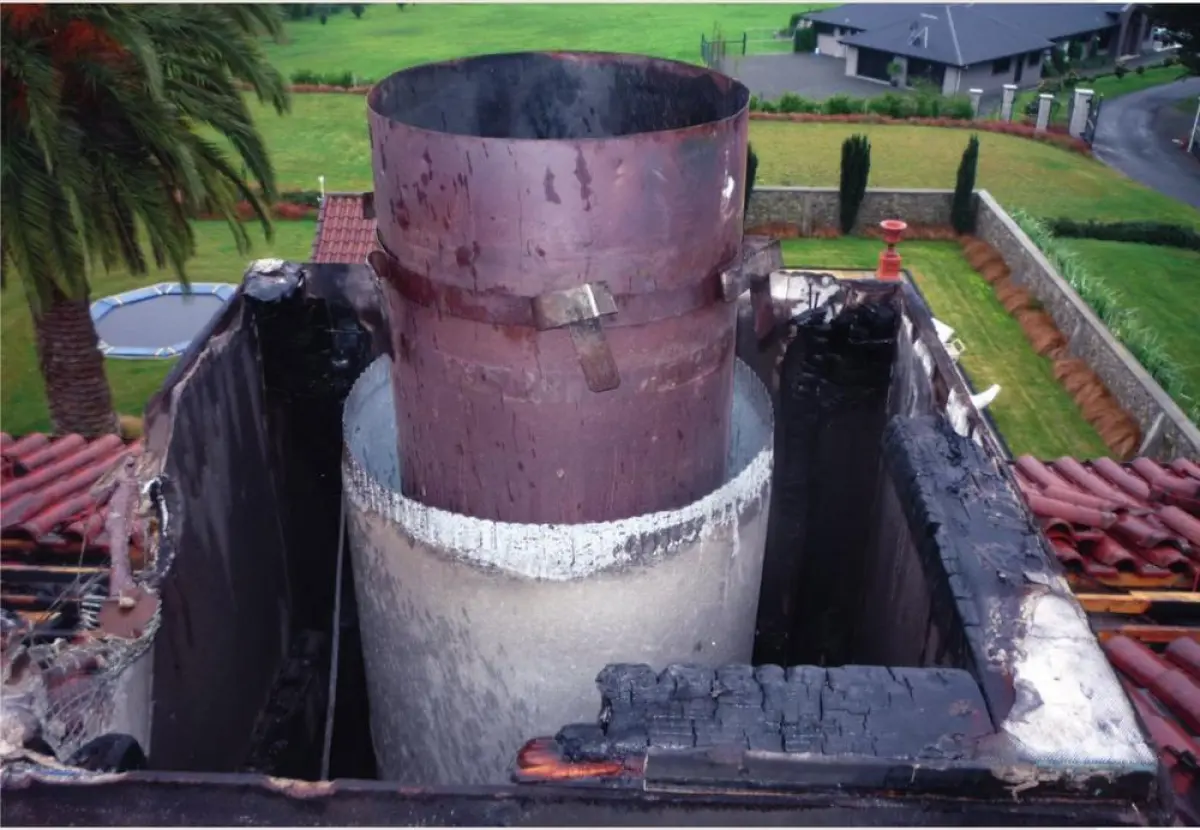 Photograph showing the top of a roof with a charred cylindrical chimney passing through a square opening, surrounded by dark flashing and roofing materials